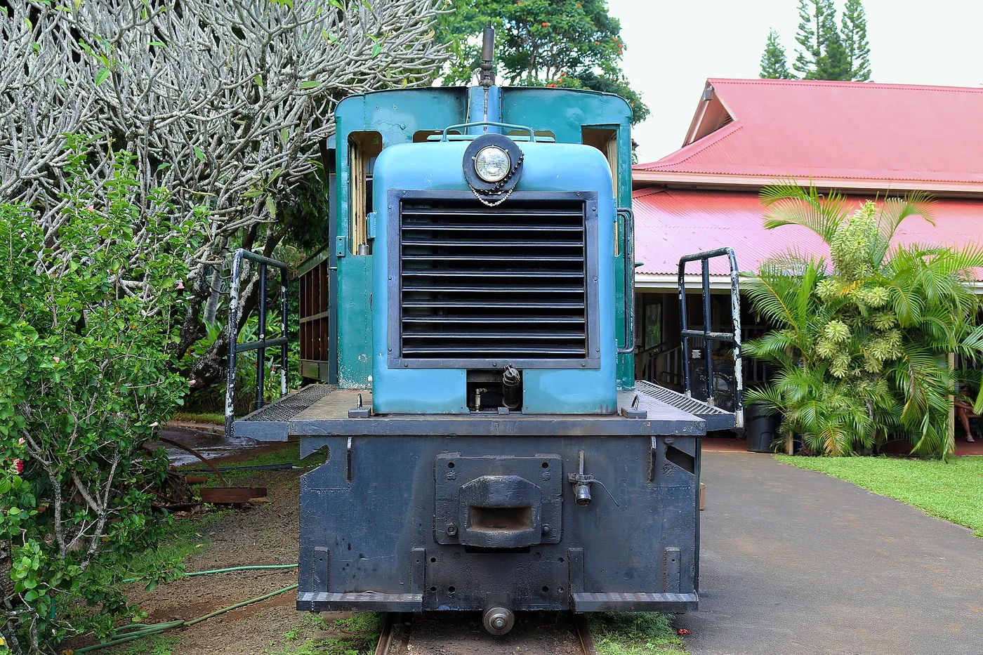 A blue vintage diesel locomotive on a short track among palms beside a red-roofed plantation building