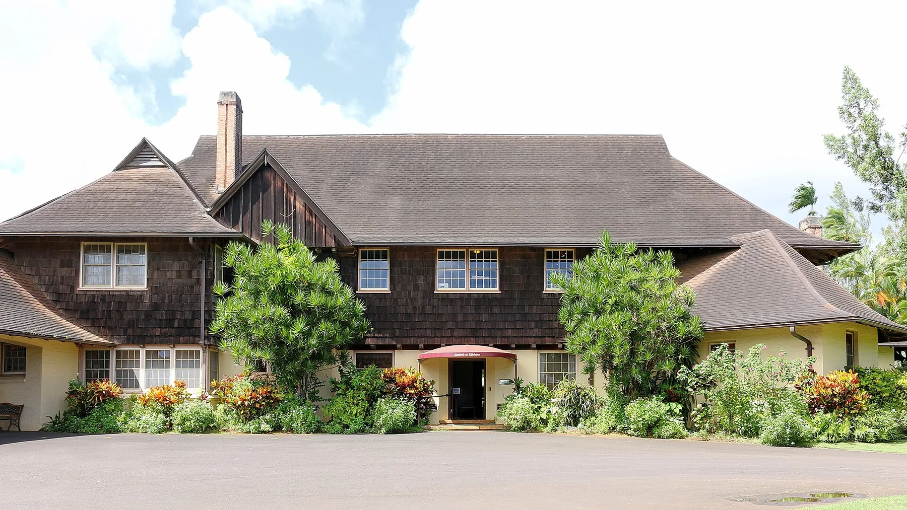 The dark-shingled two-storey manor house at Kilohana Plantation behind tropical flowerbeds and a small red entrance awning
