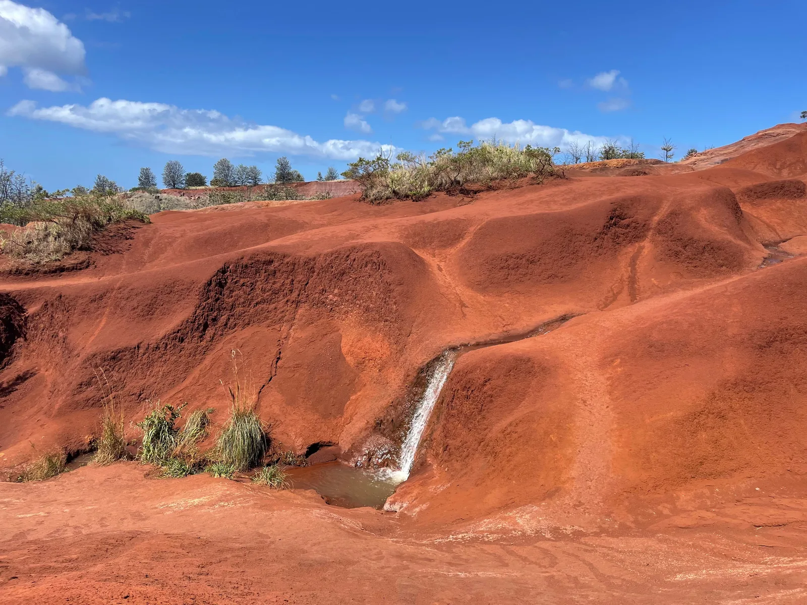 A thin waterfall running down rust-red earth in Kauaʻi under a blue sky with scattered cloud